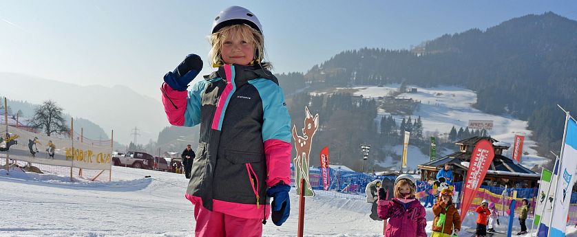 Ein kleines Kind steht auf Skiern und hält den Skistock hoch, während es auf einem Förderband im Schnee steht. Im Hintergrund sind weitere Kinder und verschneite Berge zu sehen.
