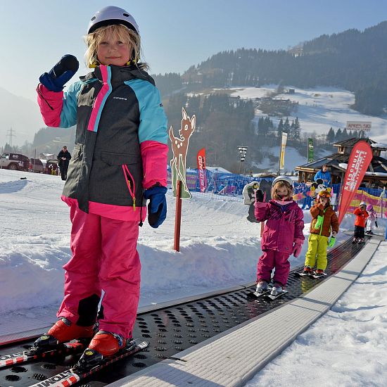 Ein kleines Kind steht auf Skiern und hält den Skistock hoch, während es auf einem Förderband im Schnee steht. Im Hintergrund sind weitere Kinder und verschneite Berge zu sehen.