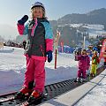 Ein kleines Kind steht auf Skiern und hält den Skistock hoch, während es auf einem Förderband im Schnee steht. Im Hintergrund sind weitere Kinder und verschneite Berge zu sehen.