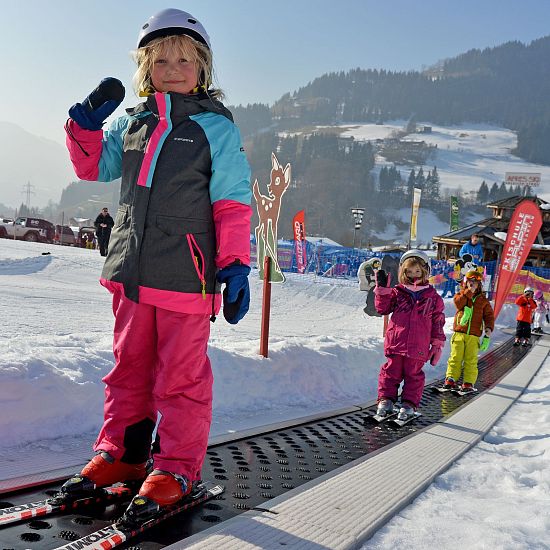 Ein kleines Kind steht auf Skiern und hält den Skistock hoch, während es auf einem Förderband im Schnee steht. Im Hintergrund sind weitere Kinder und verschneite Berge zu sehen.