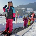 Ein kleines Kind steht auf Skiern und hält den Skistock hoch, während es auf einem Förderband im Schnee steht. Im Hintergrund sind weitere Kinder und verschneite Berge zu sehen.