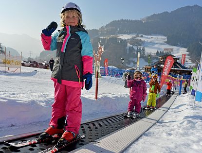 Ein kleines Kind steht auf Skiern und hält den Skistock hoch, während es auf einem Förderband im Schnee steht. Im Hintergrund sind weitere Kinder und verschneite Berge zu sehen.