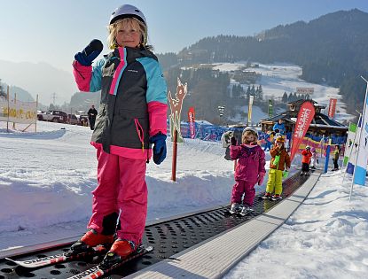 Ein kleines Kind steht auf Skiern und hält den Skistock hoch, während es auf einem Förderband im Schnee steht. Im Hintergrund sind weitere Kinder und verschneite Berge zu sehen.