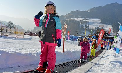 Ein kleines Kind steht auf Skiern und hält den Skistock hoch, während es auf einem Förderband im Schnee steht. Im Hintergrund sind weitere Kinder und verschneite Berge zu sehen.