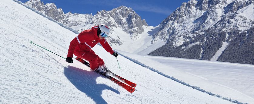 Skifahrer in roter Skiausrüstung carvt auf perfekt präparierter Piste im Skigebiet Wilder Kaiser Brixental mit verschneiten Gipfeln im Hintergrund