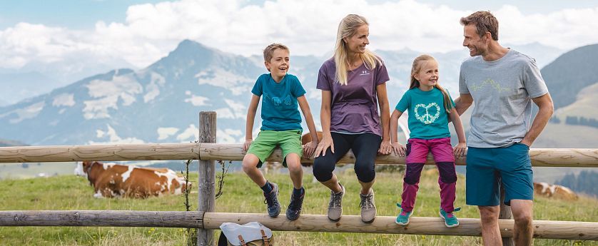 Familie beim Wandern macht eine Pause sitz auf einem Holzbalken eines Zaunes, Rucksatz steht am Boden und eine Kuh sowie Alpenlanschaft im Hintergrund zu sehen