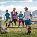 Familie beim Wandern macht eine Pause sitz auf einem Holzbalken eines Zaunes, Rucksatz steht am Boden und eine Kuh sowie Alpenlanschaft im Hintergrund zu sehen