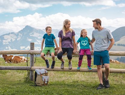 Familie beim Wandern macht eine Pause sitz auf einem Holzbalken eines Zaunes, Rucksatz steht am Boden und eine Kuh sowie Alpenlanschaft im Hintergrund zu sehen