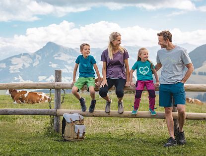 Familie beim Wandern macht eine Pause sitz auf einem Holzbalken eines Zaunes, Rucksatz steht am Boden und eine Kuh sowie Alpenlanschaft im Hintergrund zu sehen