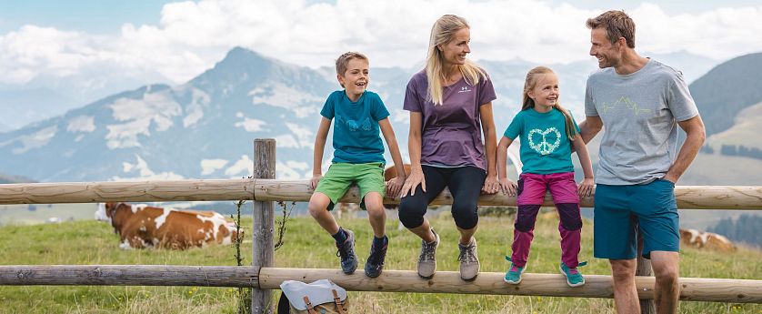 Familie beim Wandern macht eine Pause sitz auf einem Holzbalken eines Zaunes, Rucksatz steht am Boden und eine Kuh sowie Alpenlanschaft im Hintergrund zu sehen