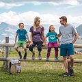 Familie beim Wandern macht eine Pause sitz auf einem Holzbalken eines Zaunes, Rucksatz steht am Boden und eine Kuh sowie Alpenlanschaft im Hintergrund zu sehen