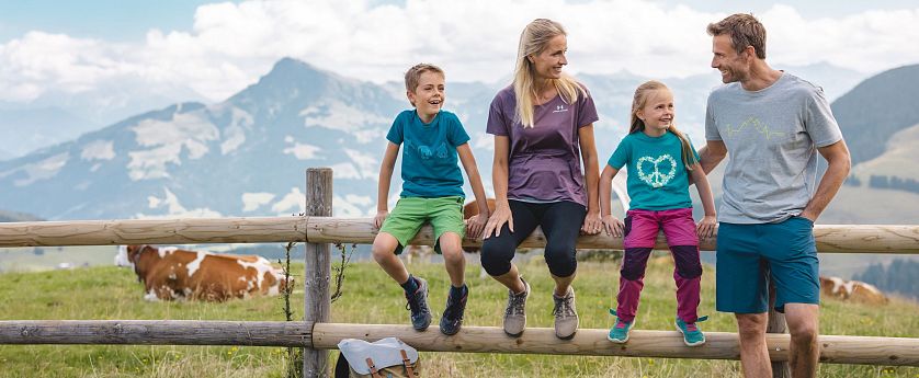 Familie beim Wandern macht eine Pause sitz auf einem Holzbalken eines Zaunes, Rucksatz steht am Boden und eine Kuh sowie Alpenlanschaft im Hintergrund zu sehen