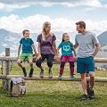 Familie beim Wandern macht eine Pause sitz auf einem Holzbalken eines Zaunes, Rucksatz steht am Boden und eine Kuh sowie Alpenlanschaft im Hintergrund zu sehen
