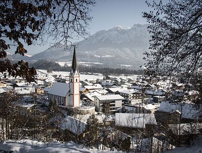 Winterliche Stadtlandschaft mit schneebedeckten Dächern und einem Kirchturm im Zentrum. Im Hintergrund sind schneebedeckte Berge sichtbar.