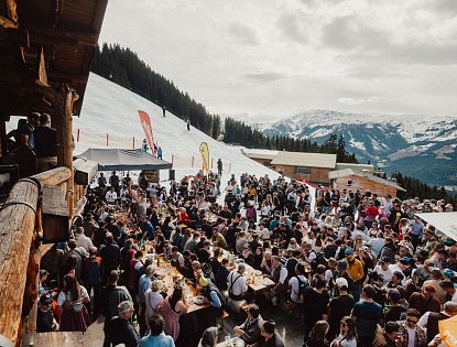 Eine lebhafte Szene in den Bergen, wo zahlreiche Menschen an langen Tischen im Freien essen und feiern. Schnee bedeckt den Boden, und die Berge sind im Hintergrund sichtbar.