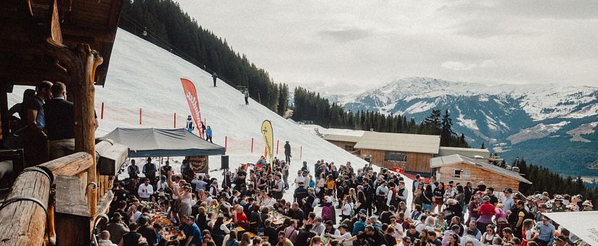 Eine lebhafte Szene in den Bergen, wo zahlreiche Menschen an langen Tischen im Freien essen und feiern. Schnee bedeckt den Boden, und die Berge sind im Hintergrund sichtbar.