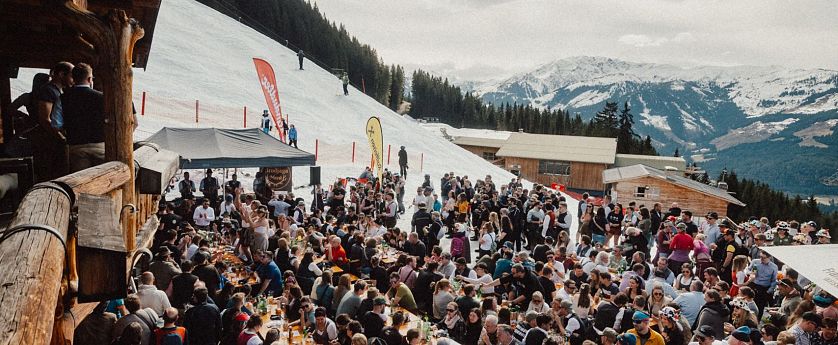 Eine lebhafte Szene in den Bergen, wo zahlreiche Menschen an langen Tischen im Freien essen und feiern. Schnee bedeckt den Boden, und die Berge sind im Hintergrund sichtbar.