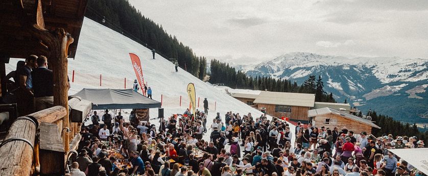 Eine lebhafte Szene in den Bergen, wo zahlreiche Menschen an langen Tischen im Freien essen und feiern. Schnee bedeckt den Boden, und die Berge sind im Hintergrund sichtbar.