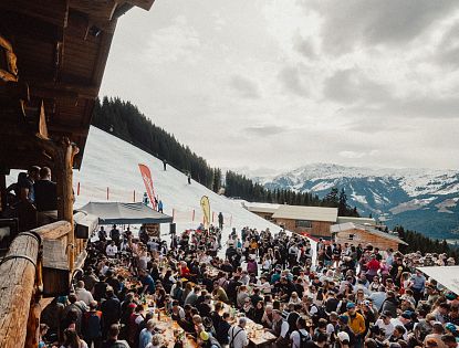 Eine lebhafte Szene in den Bergen, wo zahlreiche Menschen an langen Tischen im Freien essen und feiern. Schnee bedeckt den Boden, und die Berge sind im Hintergrund sichtbar.