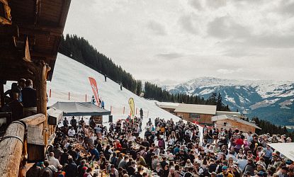 Eine lebhafte Szene in den Bergen, wo zahlreiche Menschen an langen Tischen im Freien essen und feiern. Schnee bedeckt den Boden, und die Berge sind im Hintergrund sichtbar.