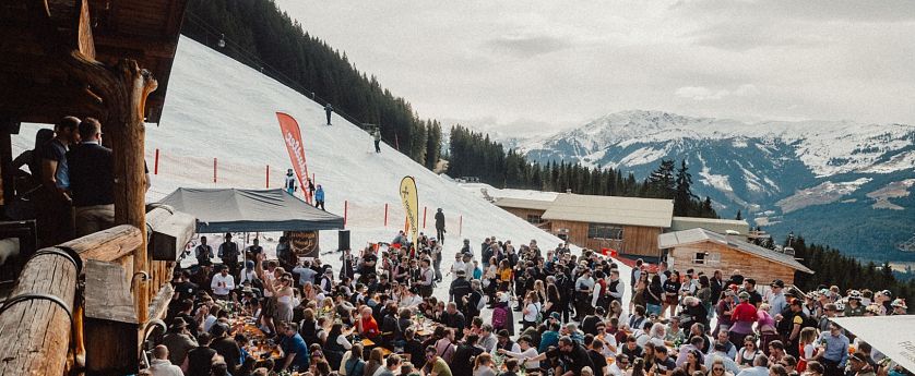 Eine lebhafte Szene in den Bergen, wo zahlreiche Menschen an langen Tischen im Freien essen und feiern. Schnee bedeckt den Boden, und die Berge sind im Hintergrund sichtbar.