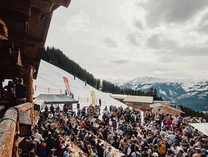 Eine lebhafte Szene in den Bergen, wo zahlreiche Menschen an langen Tischen im Freien essen und feiern. Schnee bedeckt den Boden, und die Berge sind im Hintergrund sichtbar.