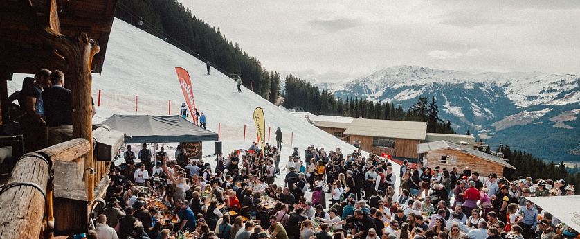 Eine lebhafte Szene in den Bergen, wo zahlreiche Menschen an langen Tischen im Freien essen und feiern. Schnee bedeckt den Boden, und die Berge sind im Hintergrund sichtbar.