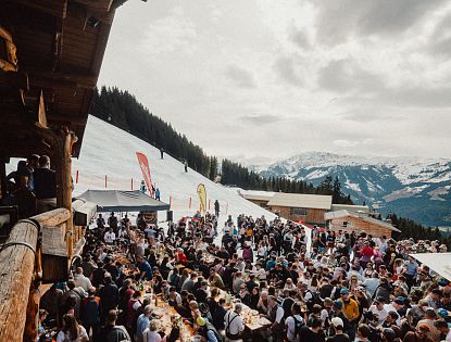 Eine lebhafte Szene in den Bergen, wo zahlreiche Menschen an langen Tischen im Freien essen und feiern. Schnee bedeckt den Boden, und die Berge sind im Hintergrund sichtbar.