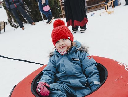 Ein fröhliches Kind in Winterkleidung, das auf einem Schlitten im Schnee sitzt, trägt eine rote Mütze und schaut in die Kamera.