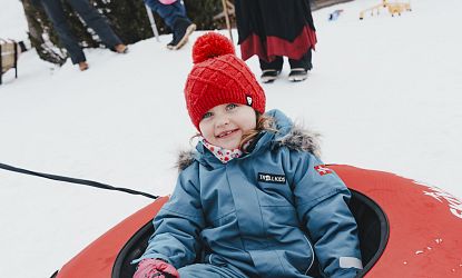 Ein fröhliches Kind in Winterkleidung, das auf einem Schlitten im Schnee sitzt, trägt eine rote Mütze und schaut in die Kamera.