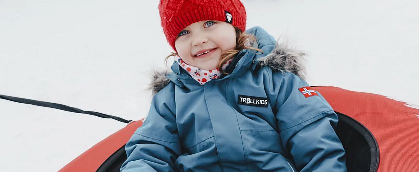Ein Kind mit roter Mütze und blauer Jacke sitzt lächelnd in einem Schlitten auf Schnee.