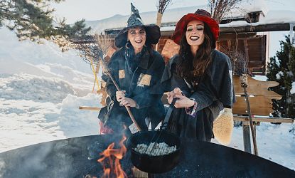 Twee vrouwen in heksenkostuums koken boven een vuur in een sneeuwlandschap, met lachende gezichten en kleurrijke hoeden.