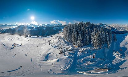 Eine weitläufige Winterlandschaft zeigt schneebedeckte Berge, einen klaren blauen Himmel und einen Wald aus hohen, mit Schnee bedeckten Bäumen. Pisten und Lifte sind sichtbar.