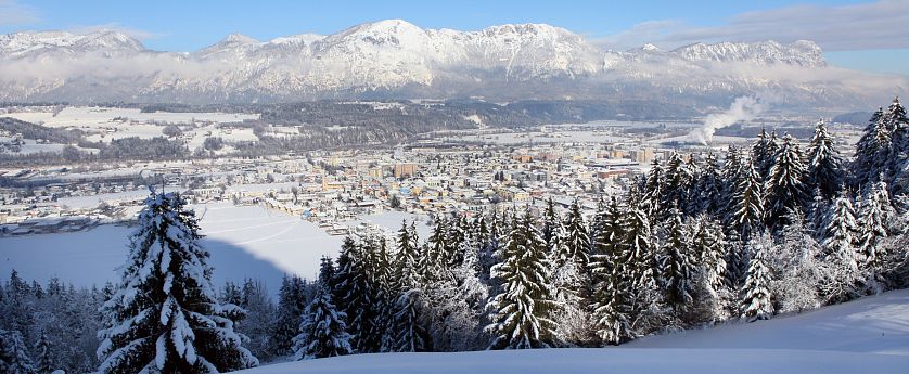 Winterlandschaft mit schneebedeckten Bäumen im Vordergrund und einer Stadt im Tal. Im Hintergrund erhebt sich eine Bergkette unter blauem Himmel.