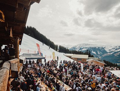 Eine lebhafte Szene in den Bergen, wo zahlreiche Menschen an langen Tischen im Freien essen und feiern. Schnee bedeckt den Boden, und die Berge sind im Hintergrund sichtbar.