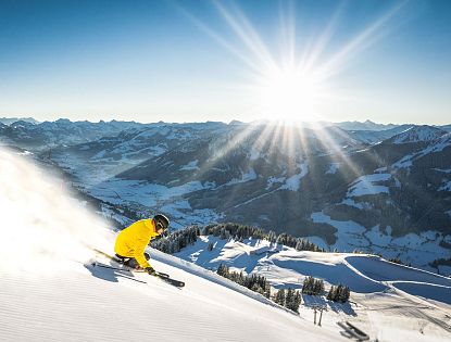 Skifahrer in gelber Jacke fährt bei Sonnenschein eine frisch präparierte Piste in der SkiWelt Wilder Kaiser Brixental mit Panoramablick auf die verschneiten Tiroler Alpen