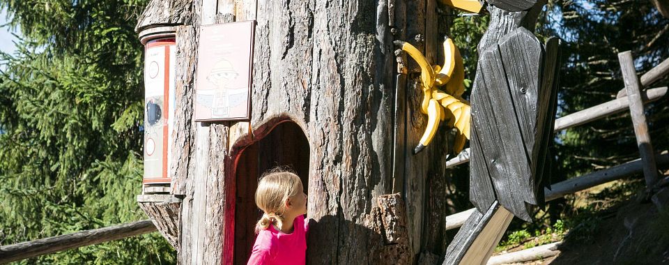 Ein Kind mit blonden Haaren in pinkem Oberteil schaut neugierig in eine Öffnung in einem großen Baumstamm auf einem Spielplatz. Es ist sonnig.