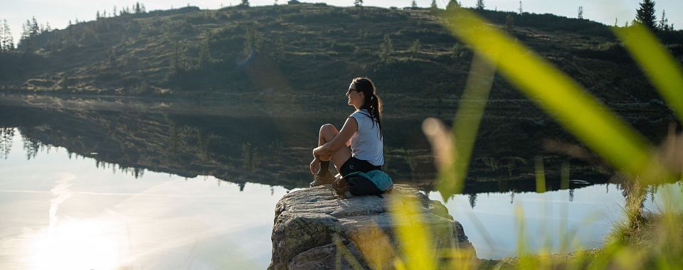 Eine Person sitzt auf einem Felsen am ruhigen See, umgeben von grünen Hügeln unter klarem Himmel. Die Sonne scheint und spiegelt sich im Wasser.