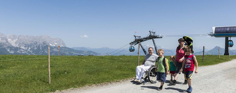 Eine Familie mit einem Rollstuhlfahrer spaziert auf einem sonnigen Weg in den Bergen. Im Hintergrund sind grüne Wiesen, ein Sessellift und ein blauer Himmel zu sehen.