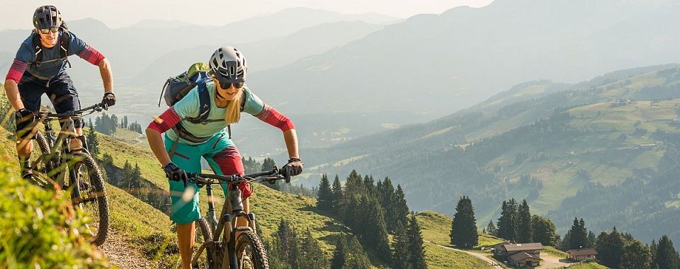 Frau mit Helm und Outdoor-Ausrüstung fährt Mountainbike auf einem Bergpfad, umgeben von Wald und Berglandschaft unter blauem Himmel.
