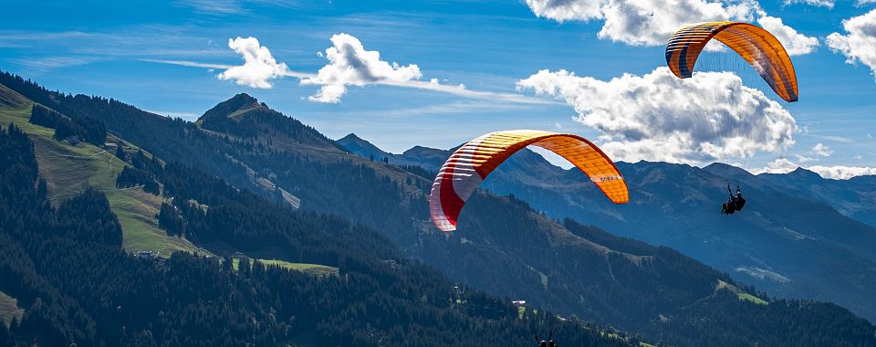 Bunte Paraglider fliegen über eine alpine Berglandschaft mit klarer Sicht auf bewaldete Hügel und kahle Gipfel unter einem blauen Himmel mit Wolken.
