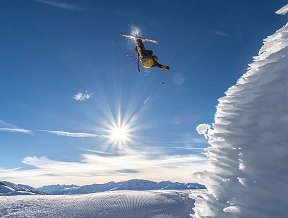 Sonniger Himmel über schneebedeckten Bergen. Die Sonne strahlt intensiv, während Wolken den blauen Himmel schmücken. Im Vordergrund ragt eine weiße Schneestruktur empor.