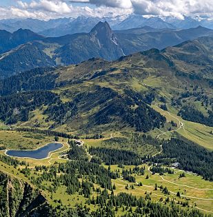 Luftaufnahme einer grünen Berglandschaft mit sanften Hügeln, Wanderwegen und verstreutem Baumbestand. Kleine Gebäude sind in der Landschaft sichtbar.