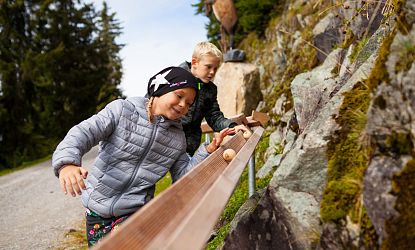 Zwei Kinder spielen fröhlich an einer Holzkugelbahn am Berghang. Sie tragen Jacken, und im Hintergrund sind Bäume und Felsen zu sehen. Die Szene wirkt lebendig.