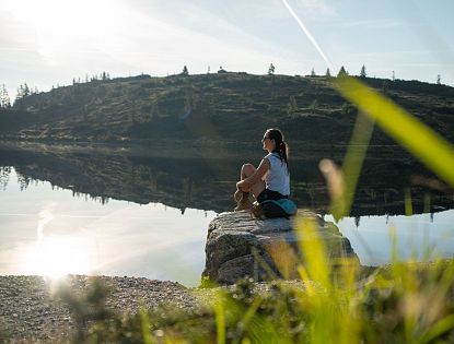Woman sitting by a calm lake, reflecting hills and sky, under a clear blue sky with greenery in the foreground.