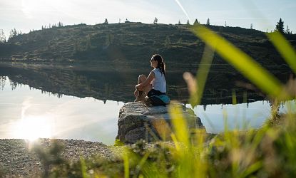 Woman sitting by a calm lake, reflecting hills and sky, under a clear blue sky with greenery in the foreground.