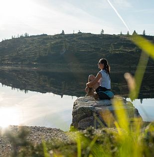 Eine Person sitzt auf einem Felsen am ruhigen See, umgeben von grünen Hügeln unter klarem Himmel. Die Sonne scheint und spiegelt sich im Wasser.