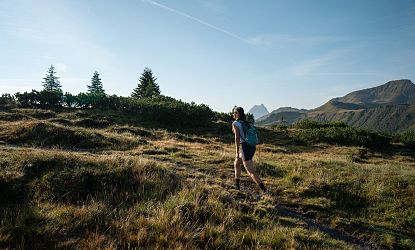 A person hikes on a grassy path with mountains in the background under a clear blue sky.