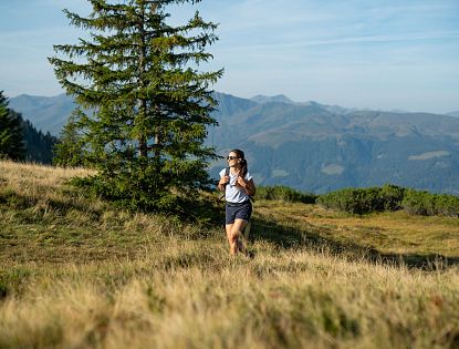 Frau joggt durch eine Berglandschaft mit Nadelbäumen und blauen Bergen im Hintergrund an einem sonnigen Tag.