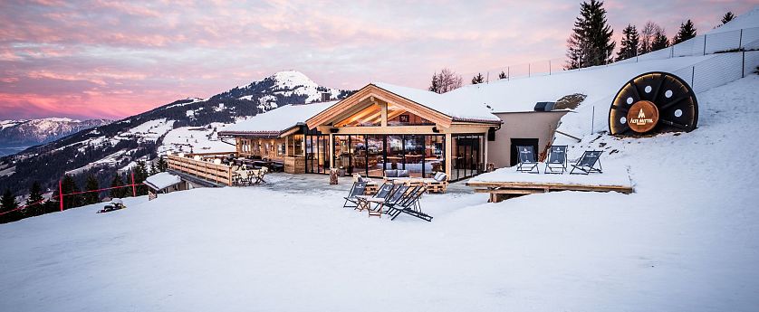 Een moderne berghut met grote ramen en sneeuw. Ligstoelen staan buiten op het terras met een berg op de achtergrond onder een lichtroze hemel.